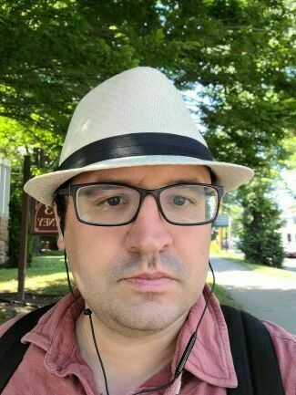This close-up photograph features a man with dark-rimmed glasses wearing a light-colored fedora with a black band. He is looking directly at the camera with a neutral expression against a brightly lit, blurred outdoor background with trees and a sign. The image captures him from the shoulders up, highlighting his accessories and the sunny, daytime setting.