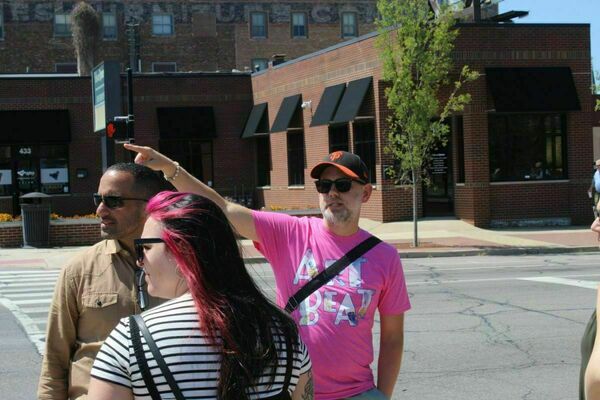 Carl Hetler, the City of South Bend's Director of Unsheltered gives a tour of downtown South Bend. He is wearing a sunglasses points across a street while standing with a group of people.