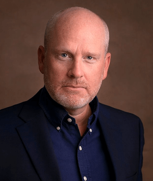 A close-up image of Charles Powell, a bald man with a reddish-blond beard, wearing a navy-blue suit with a dark blue shirt. The background is a light brown.