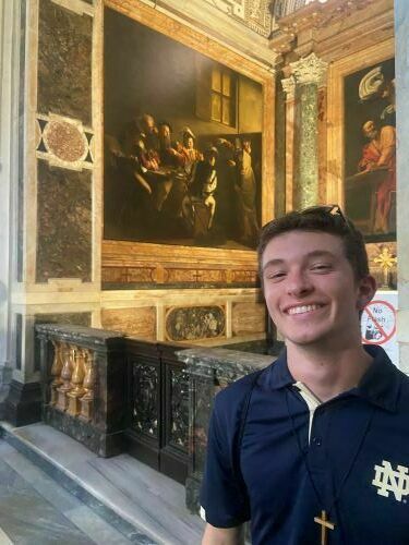 A young man, Nicholas Tarini, stands smiling indoors in front of a large religious painting and ornate walls.
