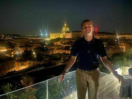Nicholas Tarini stands at night on a balcony overlooking a brightly lit cityscape. Domed buildings and towers are visible in the distance.