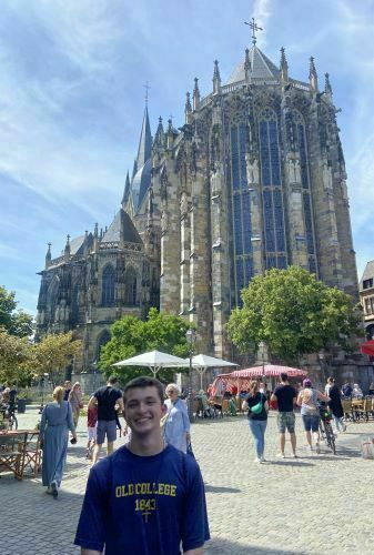 Nicholas Tarini stands outdoors in a public square wearing a blue “College Leisure” t-shirt. Behind him is a large Gothic-style cathedral with trees and people around.