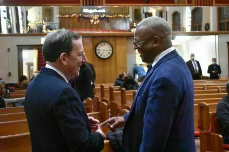 Dr. Rev. Michael Nabors, wearing glasses and a suit, shakes hands with another man inside a church sanctuary. Wooden pews and a large clock are visible in the background.