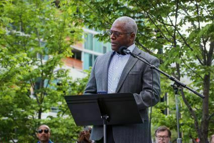 A man, identified as Dr. Rev. Michael Nabors, stands outdoors at a podium with a microphone, delivering a speech. Trees and a few seated listeners are visible in the background.