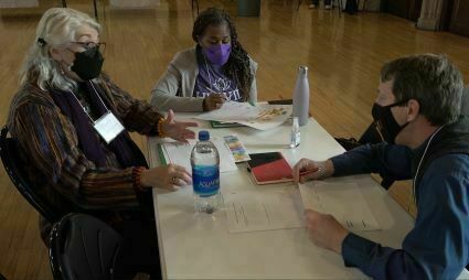 Three people sit around a rectangular table engaged in discussion. Two of them wear face masks. Papers, notebooks, and a water bottle are spread on the table.