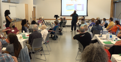 Second Baptist Church Eliminating Racism.
A speaker addresses a room of seated attendees. Round tables are arranged throughout the room, each with multiple participants facing the front where a presentation is being displayed.