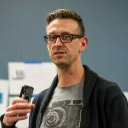 A photograph of Roman Williams speaking at an event. He is a man with short, dark blond hair and glasses, wearing a black graphic T-shirt with a gray cardigan over it. He holds a microphone and appears to be speaking in front of a whiteboard covered with sticky notes and printed materials
