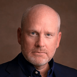A close-up image of Charles Powell, a bald man with a reddish-blond beard, wearing a navy-blue suit with a dark blue shirt. The background is a light brown.