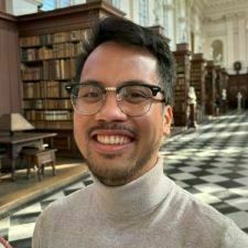 Headshot of an Asian man with dark hair, glasses, and a light extended goatee. He is smiling, wears a light colored turtleneck, and has on glasses.