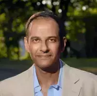 Headshot of Moustafa Bayoumi who wears a tan jacket with a blue shirt. The top button is open. He look directly into the camera with a slight smile.
