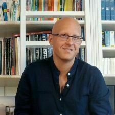 Portrait of a man in front of a bookcase, He is wearing a blue collared shirt unbuttoned at the top. He is earing glasses and smiling at the camera.