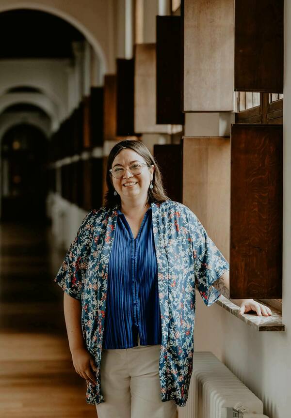 A picture of a woman with long dark hair, glasses, and a blue shirt with flowered overlay. The woman is standing with her hand resting on a sill.