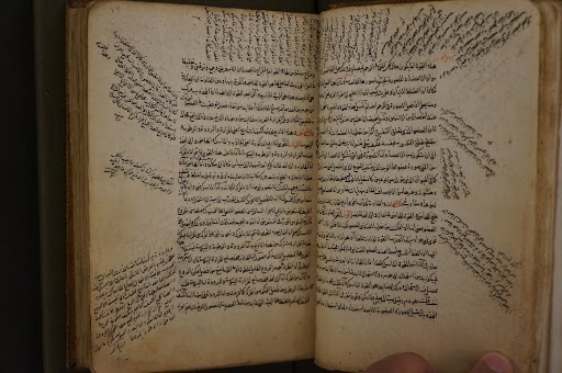 This image features an open, aged manuscript of Ibn al-Mubarak al-Qazvini's Sharh al-Mujaz, showing two pages of dense Arabic calligraphy. The text is characterized by its main body of prose supplemented by extensive marginalia, with handwritten notes curving and slanting around the primary columns.