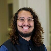 This headshot features a smiling man with long, curly brown hair and a distinctive, well-groomed handlebar mustache and beard. He is wearing round glasses and a dark, high-collared vest over a blue shirt, set against a softly blurred indoor background.