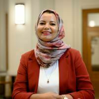 Gemini said
The image features a smiling woman wearing a patterned pink and grey hijab and a burnt orange blazer over a white top. She is posed in a professional indoor setting with soft lighting and a blurred background that highlights her friendly expression.
