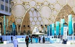 This photograph captures the Al Wasl Dome at Expo City Dubai, adorned with vibrant teal banners for the COP28 UN Climate Change Conference. Several attendees are seen walking across the patterned plaza toward the entrance of the massive, intricate lattice structure.