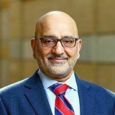 The image features a man with glasses and a graying beard, dressed in a professional navy blue blazer and a red and blue striped tie. He is smiling warmly against a soft-focus architectural background, conveying a friendly yet distinguished presence.