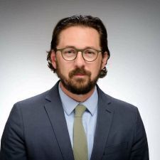 This studio headshot features a man with dark hair and a full beard, wearing olive-toned glasses that match his slim tie. He is dressed in a classic navy blue blazer and a light blue dress shirt, posed against a clean, gradient grey background.