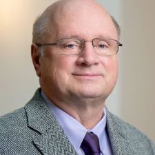 This headshot features an older man with a kind expression and wire-rimmed glasses, dressed professionally in a textured grey blazer and a purple tie. He is positioned against a neutral, softly lit background that gives the portrait a clean and approachable feel.
