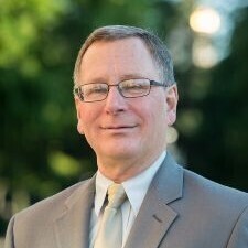 This is a professional headshot of a man with short, brown hair and glasses, posing with a calm and pleasant expression. He is wearing a light gray suit jacket paired with a white dress shirt and a pale gold tie, set against a softly blurred outdoor background of green trees.