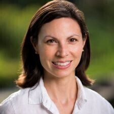 This is a professional headshot of a woman with shoulder-length dark hair, featuring a bright and approachable smile. She is wearing a crisp white button-down shirt and small stud earrings, set against a vibrant, out-of-focus background of green foliage.