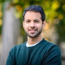 This is a portrait of a man with short, dark hair and a neatly trimmed beard, looking directly at the camera with a soft smile. He is wearing a textured, dark green crewneck sweater over a white undershirt, set against a blurred background of green and yellow foliage.