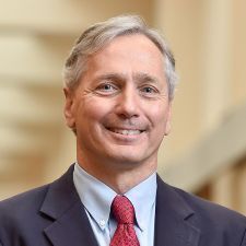 This is a headshot of a middle-aged man with short, graying hair and blue eyes, captured with a pleasant and friendly expression. He is wearing a professional ensemble consisting of a dark navy suit jacket, a light blue button-down shirt, and a patterned red necktie.