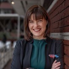 This is a professional portrait of a woman with shoulder-length brown hair and bangs, who is smiling warmly at the camera. She is dressed in a dark blazer over a teal blouse and is leaning against a textured brick wall in an outdoor setting.
