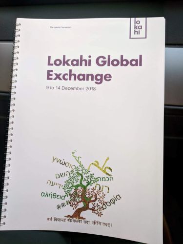 In the image, a white, spiral-bound booklet features the prominent purple title "Lokahi Global Exchange" alongside the dates "9 to 14 December 2018." The bottom of the cover is decorated with an illustration of a tree whose branches and leaves are formed by words written in various global scripts and languages.