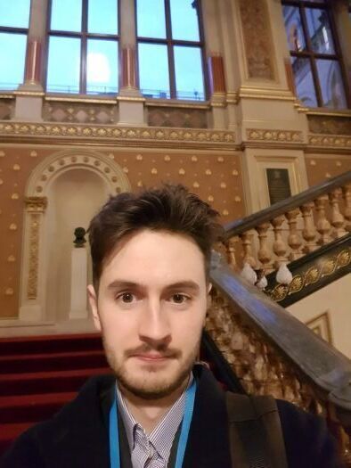 In the image, a man with a light beard and dark hair takes a close-up selfie while wearing a blue lanyard. He is standing in front of an ornate, grand staircase with red carpeting, surrounded by classical architecture featuring high arched windows and decorative wall patterns.