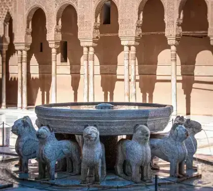 A detailed, close-up shot captures the iconic Fountain of the Lions in the foreground, showcasing its large, intricately carved stone basin supported by a ring of stylized stone lion sculptures that spout water, while the background features the courtyard's elegant, sun-drenched stucco arches and slender columns casting deep shadows.