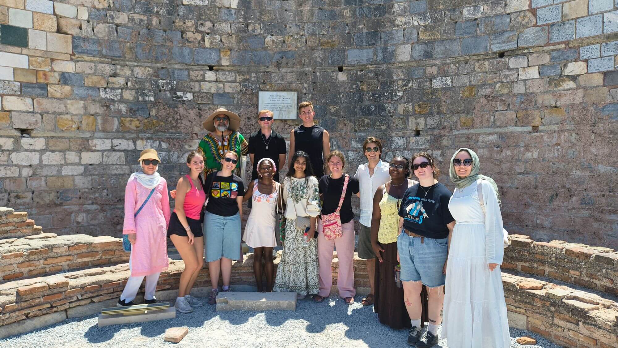 This medium wide, eye-level shot captures a group of twelve diverse people posing together against a curved, ancient stone and brick wall beneath bright sunlight, with a white informational plaque mounted directly behind them. Photo credit Mahan Mirza.