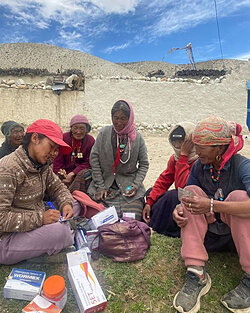Women sit on the ground with medical supplies, one is writing while others look on.